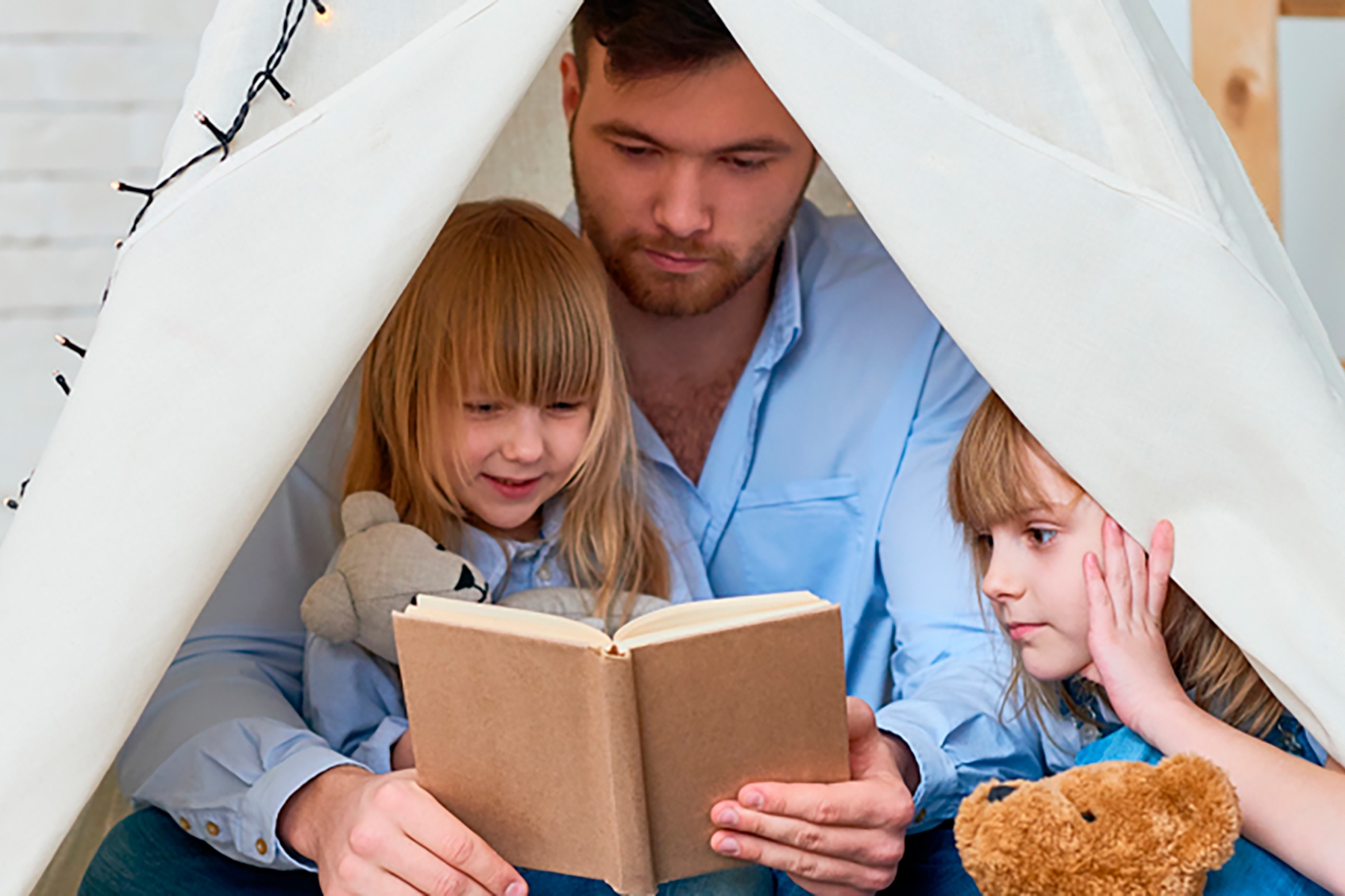 A photo of a dad sitting under an indoor canopy with his two young girls as he reads a story to them