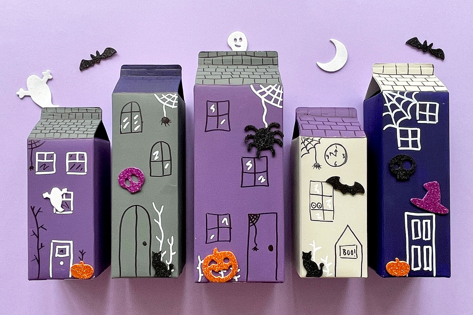 A photo of a DIY haunted village made out of empty juice cartons on a light purple background