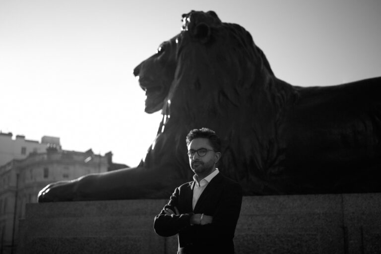 A black and white photograph of Sathnam Sanghera with his arms crossed, standing in front of a statue of a lion.