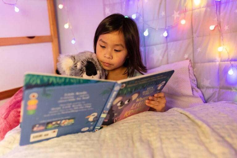 A photo of a little girl sitting in her bed reading. She is reading a bed-time story with her koala stuffed toy.